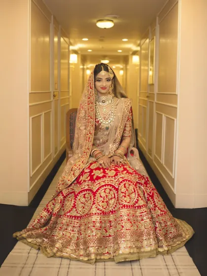 A full-length portrait of a bride in her magnificent red and gold lehenga, seated elegantly in a warmly lit corridor. This shot emphasizes bridal beauty, intricate attire, and creates a sense of regal grace.