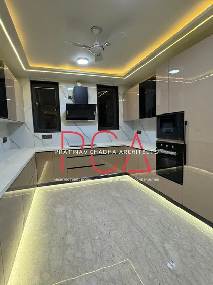 A modern, U-shaped modular kitchen from the Vasant Kunj luxury residence, designed for both functionality and aesthetic appeal. The space features high-gloss beige cabinetry, a marble-patterned backsplash, and integrated appliances, all illuminated by ambient ceiling and under-cabinet lighting.