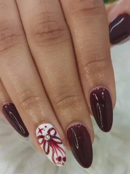 A festive and detailed design. This look combines a deep maroon polish with a hand-painted white accent nail, featuring a ribbon design and a single rhinestone for sparkle.