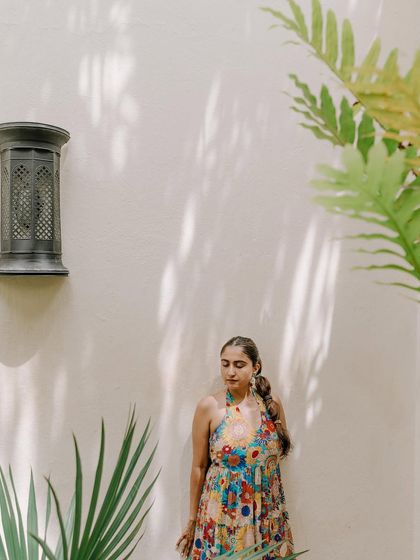 A serene portrait of the bride during her mehendi in Kenya. She stands against a white wall, framed by tropical plants and beautiful shadows, creating a calm and natural image.