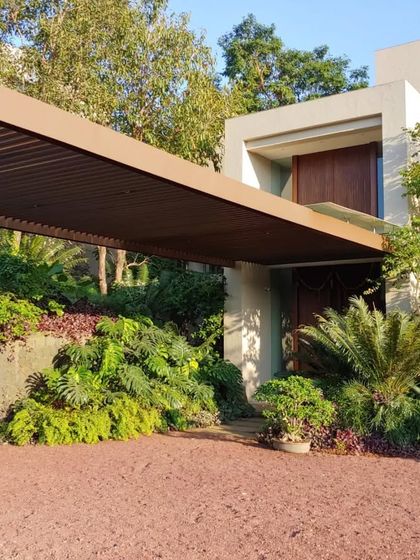 The entrance to the "House in the Hills" features a covered carport that blends into the main structure. The design uses natural earth tones and is surrounded by dense foliage to create a welcoming arrival experience.