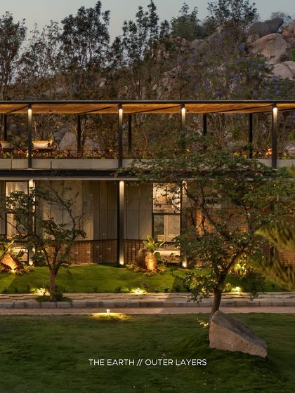 The outer layers of 'The Earth' at twilight, showing the two-story structure with its illuminated upper terrace. The soft landscape lighting highlights the trees and pathways, creating a safe and beautiful environment after dark.