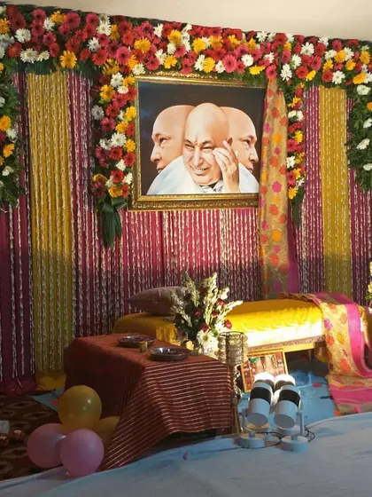 A colorful and simple stage decoration for a satsang. The backdrop uses bright yellow and pink drapes, with a large floral arrangement of gerberas and roses across the top.