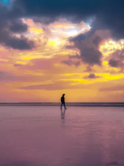 A lone figure walks on a beach under a dramatic, colorful sunset. The reflection of the sky on the wet sand creates a surreal, painterly effect, emphasizing a moment of solitary wandering.