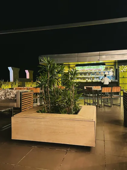 This view captures the ambiance of the rooftop cafe, with the central planter in the foreground and the glowing bar in the background. Our designs focus on creating a complete sensory experience through lighting, furniture, and plant selection.