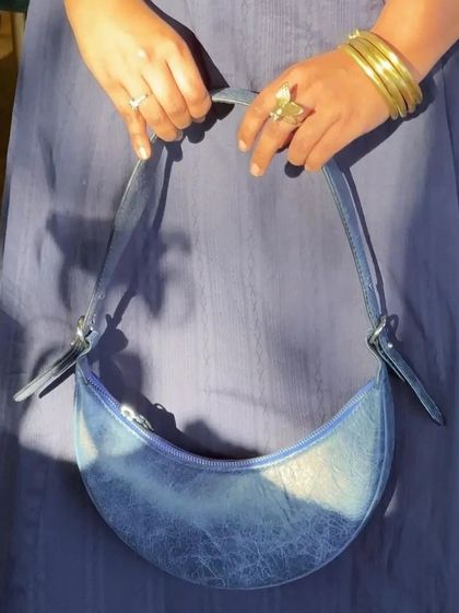 A custom Selena bag in Blue Crunch leather, held against a matching blue dress. The unique texture of the leather catches the light beautifully, highlighting the bag's distinct crescent form.