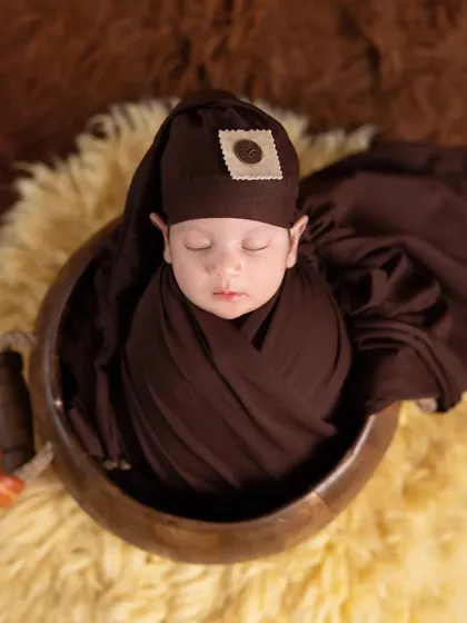 A newborn wrapped snugly in a rich, chocolate-brown swaddle. The baby rests on a soft yellow fur rug, creating a warm and cozy color combination.