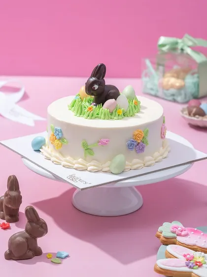 A charming Easter mini cake, decorated with a chocolate bunny sitting on green buttercream grass amidst colourful eggs. It's a delightful and festive cake for your Easter gathering.