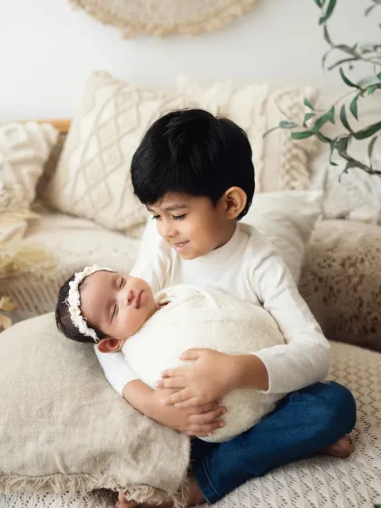 A proud big brother looks at his sleeping newborn sister, holding her gently in his lap. This image showcases a quiet, loving moment from a sibling photoshoot.