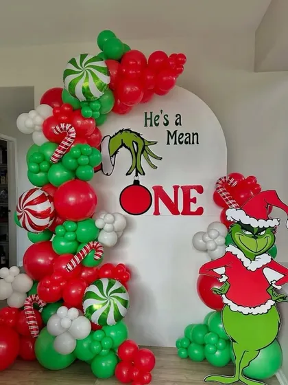 A fun and unique Christmas party theme: "He's a Mean ONE." This Grinch-themed first birthday backdrop includes a character cutout and a balloon garland in the Grinch's signature red, green, and white colors.