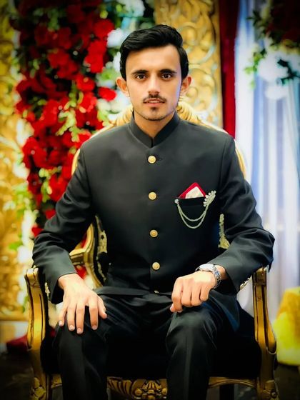 A seated portrait of a groom in a Jodhpuri suit. The red and white pocket square adds a stylish detail to his dark outfit.