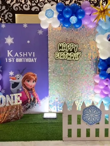 A magical first birthday with a Disney's Frozen theme, featuring a shimmer wall, character cut-outs of Anna and Elsa, and balloon pillars in icy blue, purple, and white.