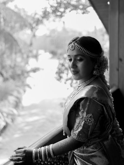 A serene black and white portrait of a South Indian bride. Her pensive look as she stands by a balcony creates a moment of quiet beauty and reflection.