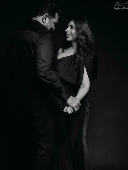 A full-length couple portrait that feels like a classic movie scene. The dark attire and deep shadows create a sense of timeless romance in this black and white photo.