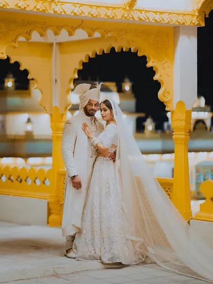 A grand, full-length portrait of a couple in matching white outfits, standing against a vibrant yellow palace backdrop at night.