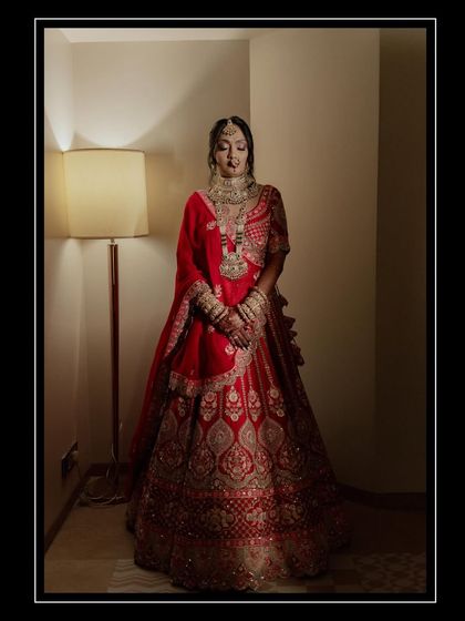 A full-length portrait of a bride in a magnificent red lehenga. The warm, dramatic lighting highlights the intricate details of her outfit and her regal presence.