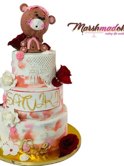This grand 3-tier, 6kg Belgian Chocolate cake was designed for a first birthday. It features a watercolour buttercream finish, edible lace, handmade flowers, and a cute, hand-sculpted teddy bear topper.