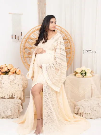 A full-length portrait of a mother-to-be in a beautiful lace dress, standing in a stylishly decorated boho corner of the studio. The look is both elegant and free-spirited.