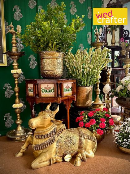 A seated brass Nandi statue surrounded by traditional lamps, wooden furniture, and lush greenery, creating a sacred and beautiful corner.