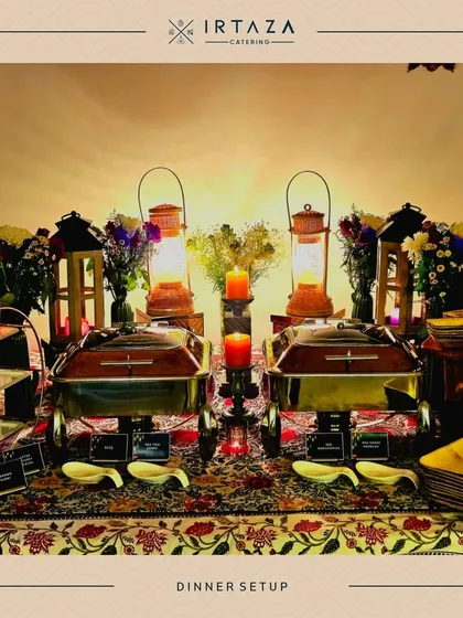 A rustic and romantic dinner setup. The table is covered with a traditional patterned cloth and lit by lanterns and candles, creating a warm and inviting atmosphere for an intimate meal.