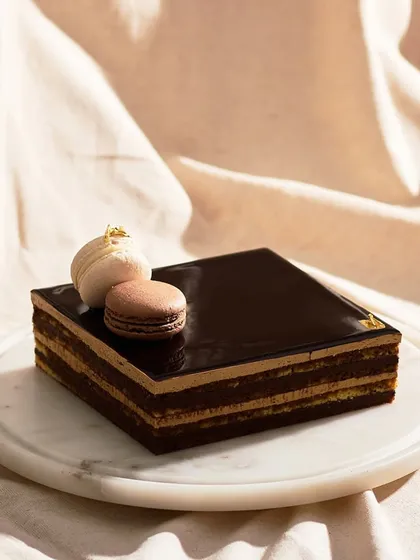 My classic Opera Cake, presented whole. Topped with two delicate macarons, its glossy chocolate glaze and clean lines are a nod to its French origins.