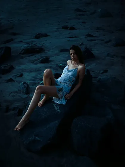 A full-length portrait of the model seated on the rocks. The deep blues of the evening light envelop the scene, creating a moody and cinematic feel.
