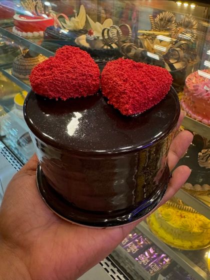 A rich, dark chocolate mini cake topped with two fuzzy red hearts. The glossy finish and simple decoration make it a sophisticated and romantic treat.