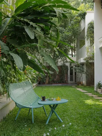 A quiet corner of a lawn with a small seating arrangement. The space is framed by flowering bushes and a stone pathway, offering a simple yet beautiful spot to enjoy the garden.