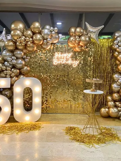 A glamorous 50th birthday setup featuring a gold sequin wall that catches the light beautifully. The scene is framed by a sophisticated balloon arch in metallic gold and silver, with large, illuminated marquee numbers proudly displaying the age.