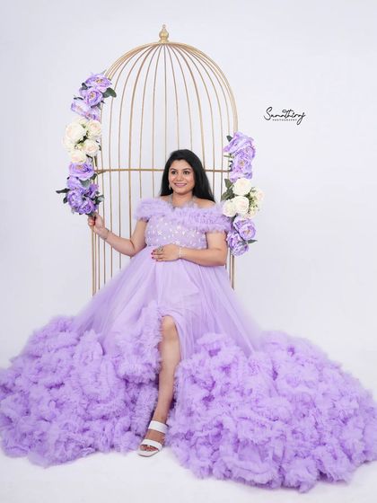 A regal portrait of a mother-to-be in a lavender gown, seated in a golden birdcage prop. The matching purple and white flowers add to the whimsical and elegant theme.