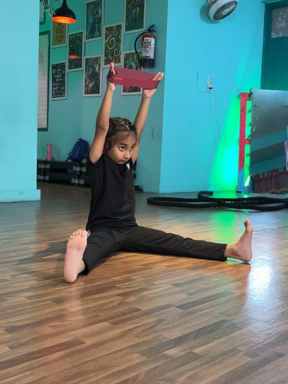 A young student demonstrates her strength and focus, holding a yoga block overhead while in a split. This exercise challenges her core stability and shoulder strength.