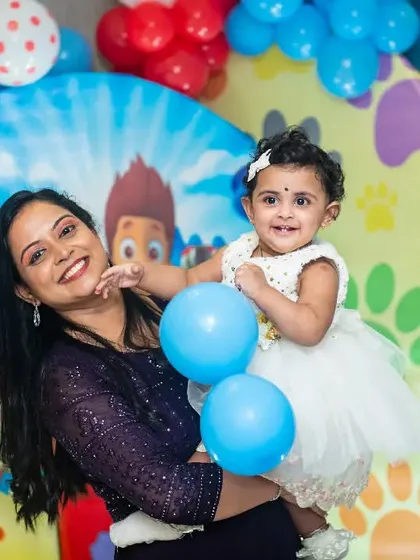 A mother holds her daughter, who is playing with a blue balloon, at her fun-filled first birthday party.