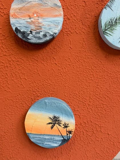 A close-up of some of the miniature travel paintings. Here you can see a sunset over the ocean and a beach with palm trees, each painted on a small wooden circle.