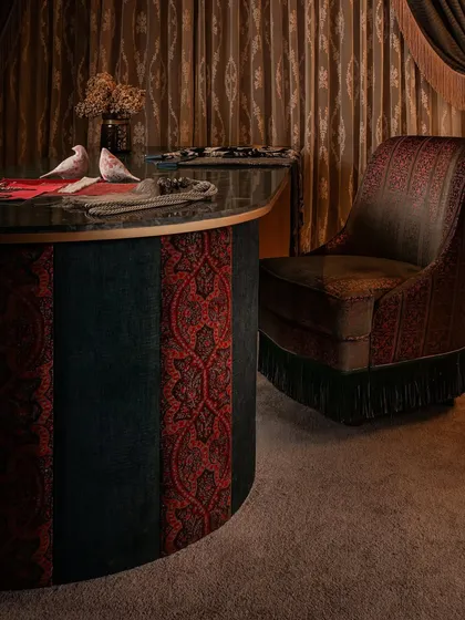 The curved client table is clad in alternating panels of rich red and deep teal fabric, creating a bold statement. The custom-embroidered swivel chair adds another layer of texture and craft, completing this maximalist composition.