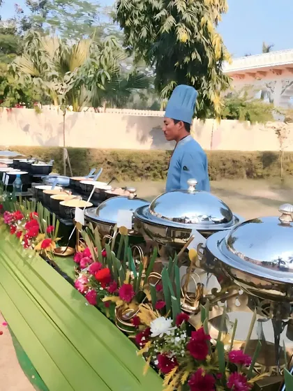An outdoor buffet line with a chef in uniform attending to the dishes. The setup features classic silver chafing dishes decorated with vibrant red and pink flowers for a daytime event.