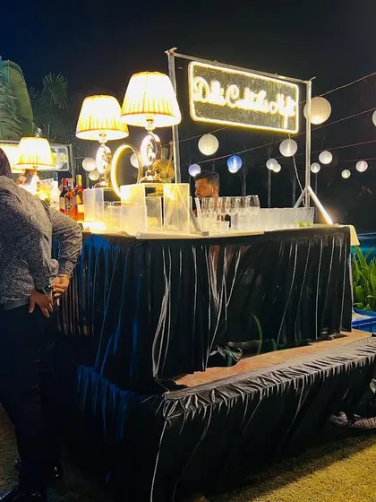 Our portable cocktail bar setup is perfect for outdoor events. This one features a "Delhi Cocktails Night" neon sign, elegant lamps, and paper lanterns hanging in the background.
