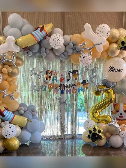 A paw-sitively adorable dog paw theme for a 2nd birthday. This setup includes a full balloon arch in gray, gold, and white, accented with bone-shaped and paw-print foil balloons, and a "Happy Birthday" banner with cute dog characters.
