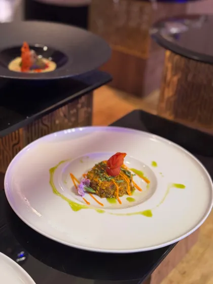A vibrant dish where a tower of grains and vegetables is crowned with a red garnish, surrounded by artistic drizzles of green oil.