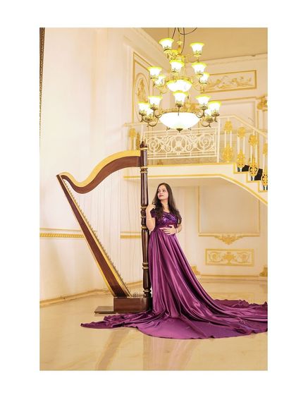 A regal solo portrait of the bride-to-be standing by a harp in a luxurious hall, her long magenta gown spread elegantly on the floor.