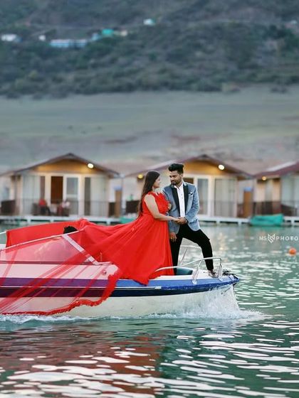 A dynamic shot of a speedboat creating a wake, with the couple enjoying the ride. The woman's red gown adds a splash of color and drama against the floating cottages of Tehri.