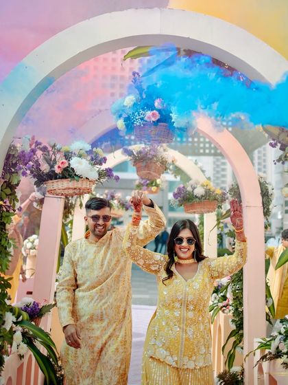 The couple's joyful entrance through a floral archway, celebrated with a burst of blue smoke, kicking off their sunset Haldi soirée.