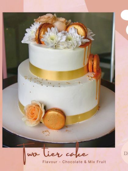 A chic two-tier cake featuring a white and gold theme. This design is elevated with a gold ribbon, macarons, and fresh flowers, perfect for an elegant celebration.