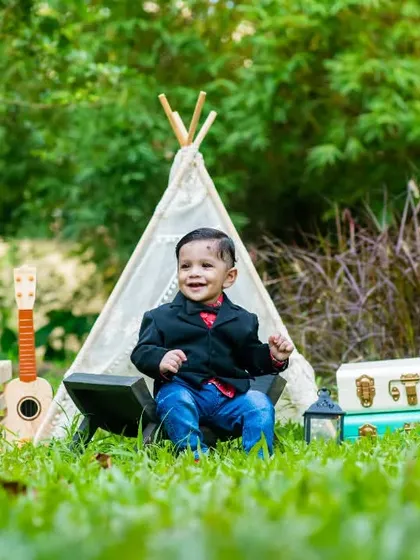 A one-year-old boy looking sharp in a little blazer, smiling for his pre-birthday photoshoot in a classic outdoor traveler theme setup.