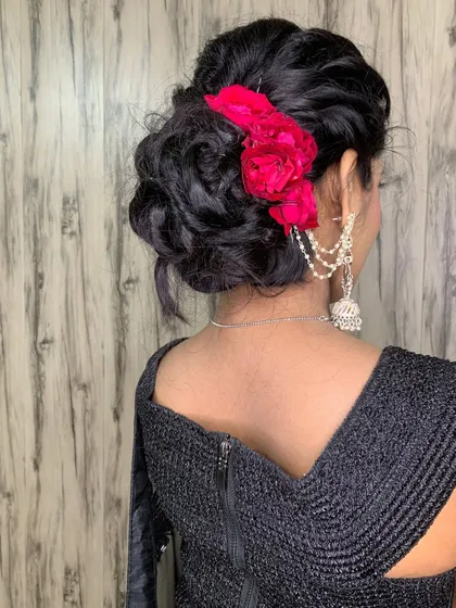 A perfect hairstyle for any wedding or festive event. This is an intricate, messy bun adorned with fresh pink flowers, combining traditional elegance with a modern touch. This kind of updo keeps hair secure while looking effortlessly beautiful.