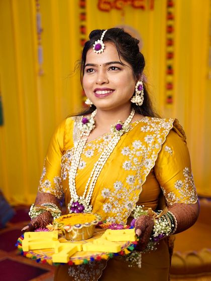 A happy Haldi bride holding a decorative plate. Her hair is styled to keep it off her face, and her floral jewelry adds a fresh, festive touch.