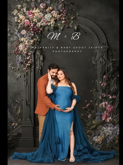 A dramatic studio portrait with a dark floral background. The couple's pose and the rich blue of the maternity gown create a look of classic elegance and romance.