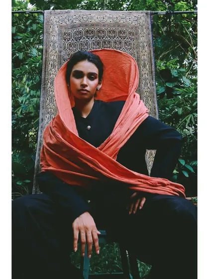 For this shot in the 'Roots' series, I styled a vibrant orange scarf as a hood against a simple black outfit. The tapestry in the background connects the outdoor setting to the indoor, cultural theme.