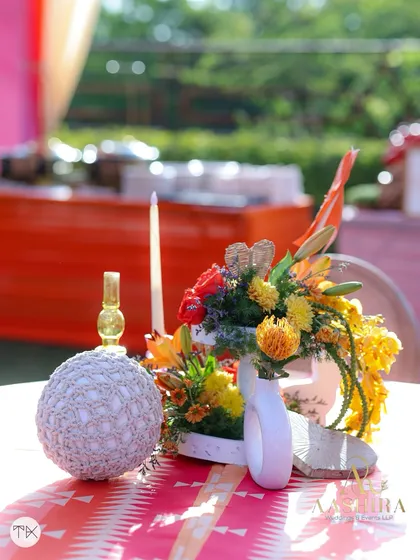 A whimsical table centerpiece for a high-tea event, featuring a mix of orange lilies, a woven ball, and a pinwheel decoration.