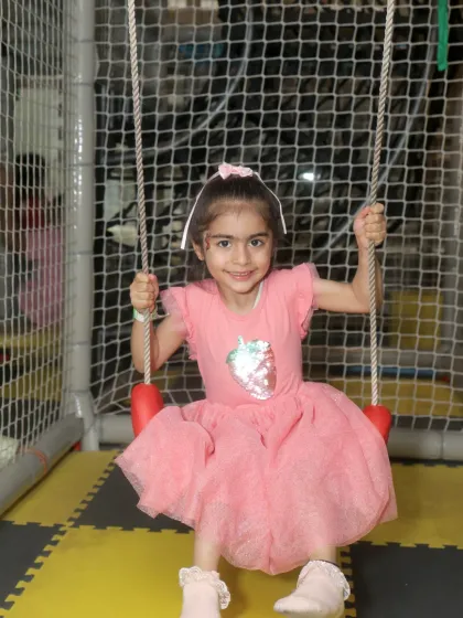 A little girl in a pink dress smiles brightly while enjoying the swing inside our netted play area.