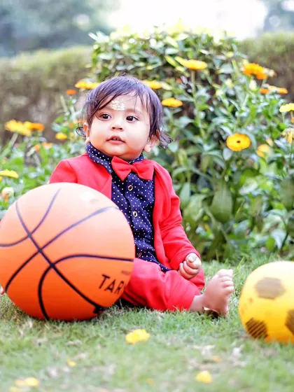 A toddler sits on the grass with a basketball and soccer ball, part of a fun, sports-themed outdoor session.